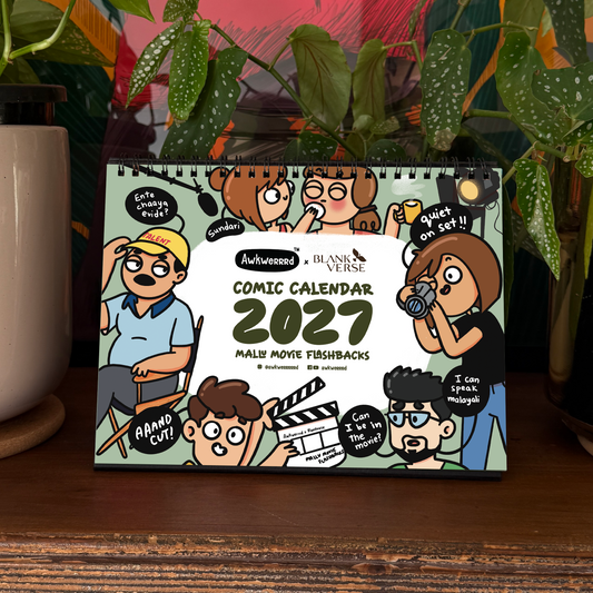 A 2027 desk calendar titled 'Comic Calendar' by Awkwerrrd is displayed on a wooden shelf in a styled home setting, surrounded by potted green plants and a colorful art print in the background.